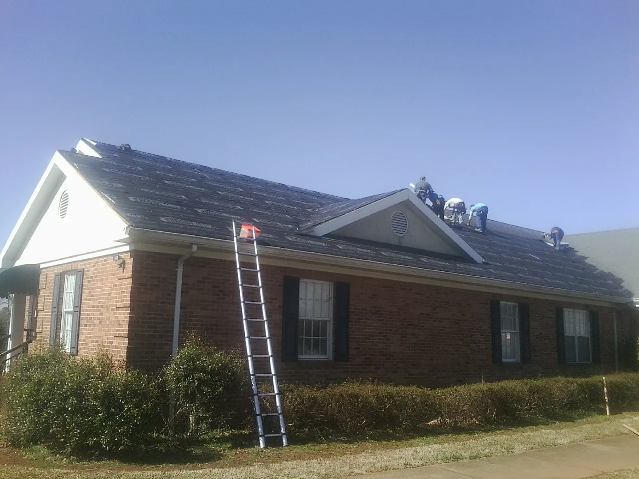 Crew tearing shingles off a brick ranch roof with a ladder set — Emergency Roof Repair work by our crew in Le Roy
